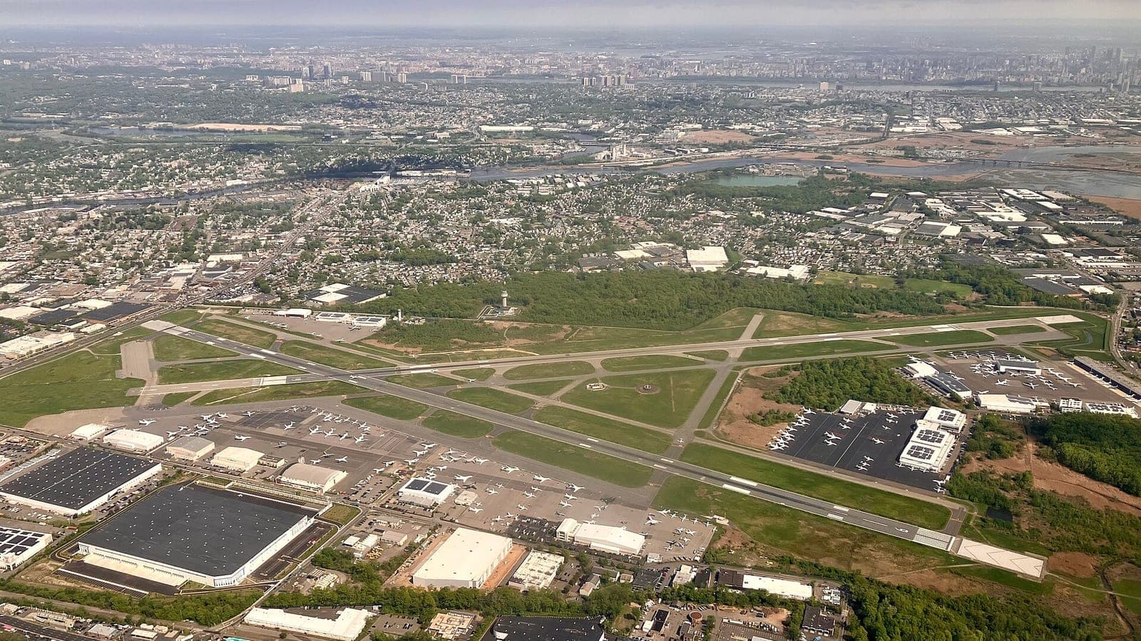 Teterboro Airport photographed from the air, its two runways crossing between rows of business jets parked on the FBO ramps, with the Manhattan skyline on the horizon.