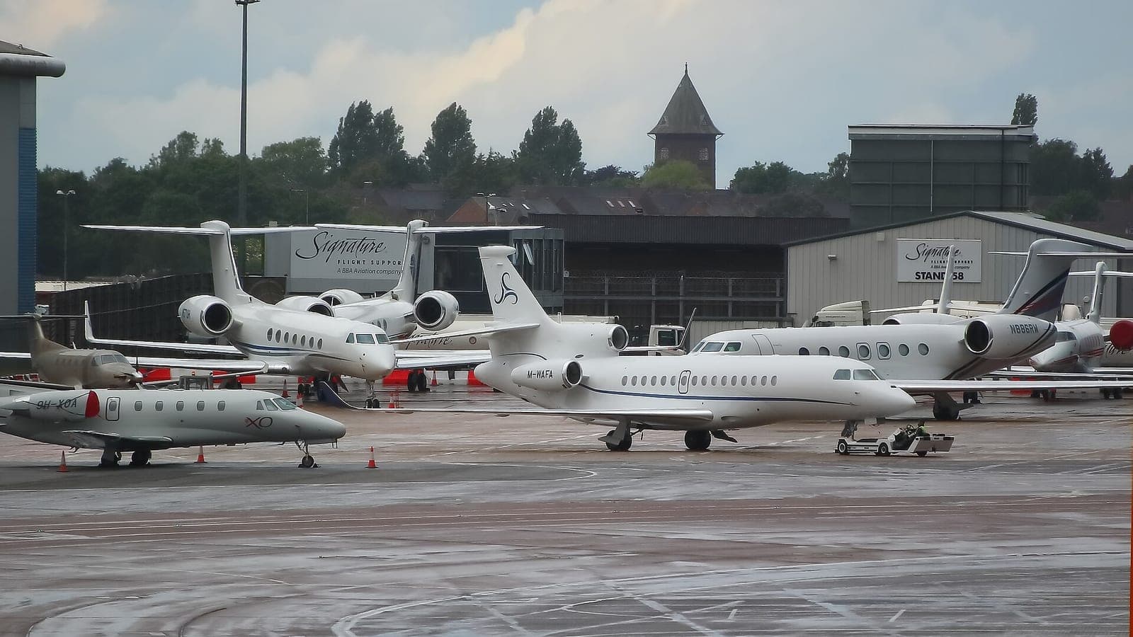 Business jets packed onto a wet apron at London Luton Airport in front of two Signature Flight Support buildings, a Citation XLS in XO livery nearest the camera and a Falcon 8X beside it being pushed back by a tug.