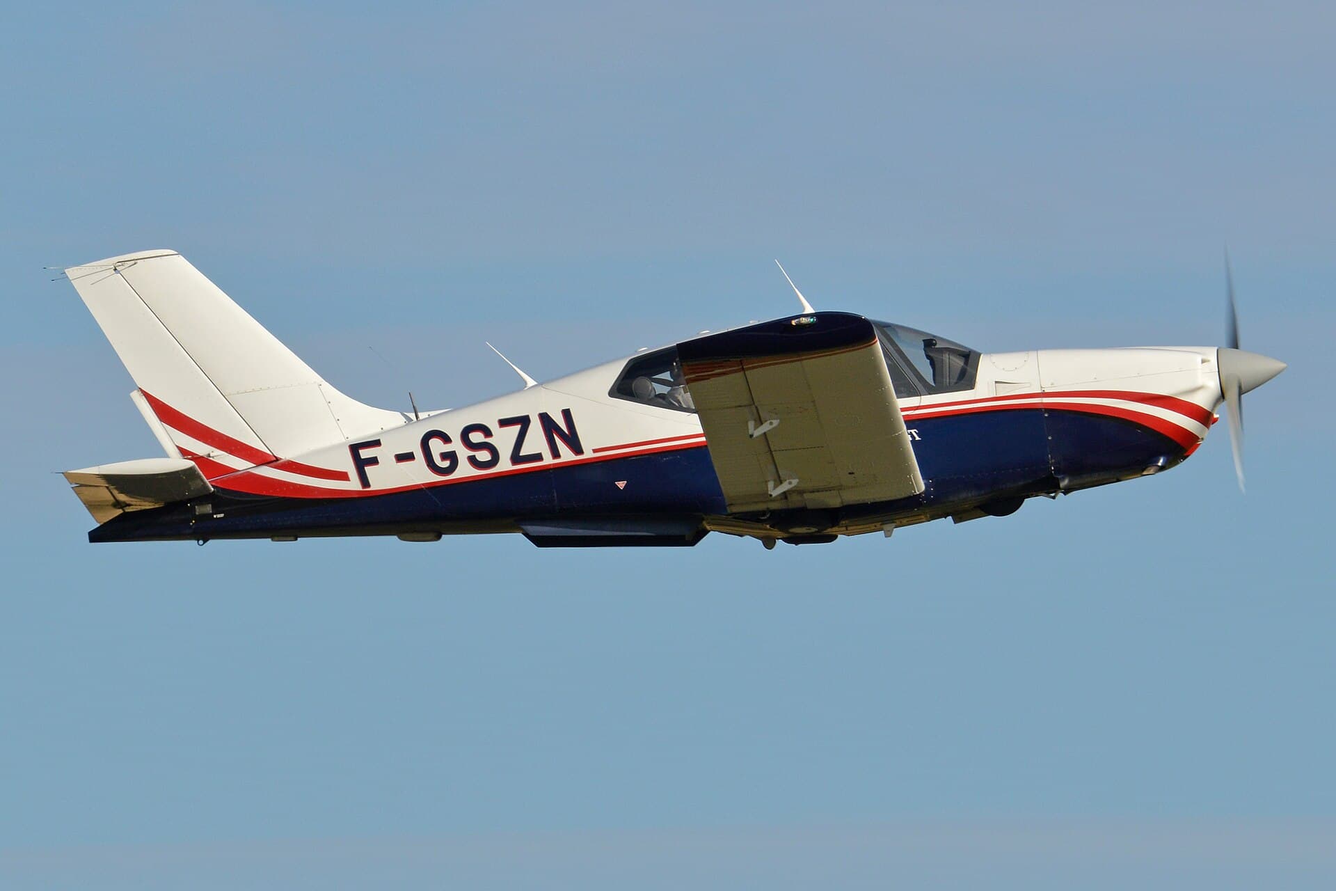 A single-engine SOCATA TB-20 Trinidad, registration F-GSZN, in a white and dark trim scheme with its low wing and distinctive gull-wing cabin doors, departing from Duxford Airfield in England on a summer evening with the landing gear still extended.