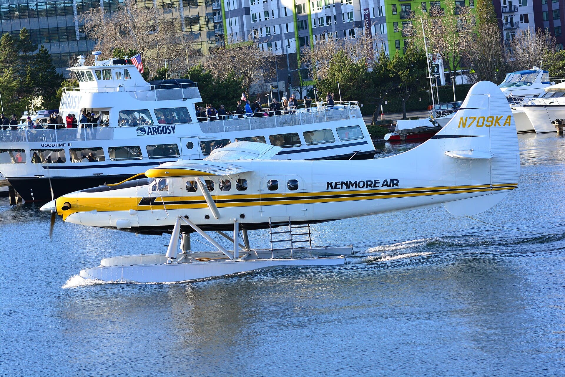 A float-equipped de Havilland Canada DHC-3 Otter in Kenmore Air markings, registration N709KA, sitting on the water at Lake Union in Seattle with a moored vessel and the lakeside waterfront behind it in flat winter daylight.