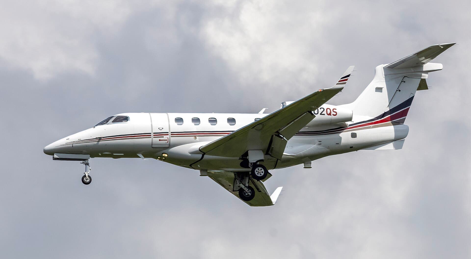 A NetJets Embraer Phenom 300, registration N302QS, on final approach with gear and flaps extended, in the operator's white fuselage and dark blue tail livery, photographed against a clear sky at Washington Dulles International Airport.
