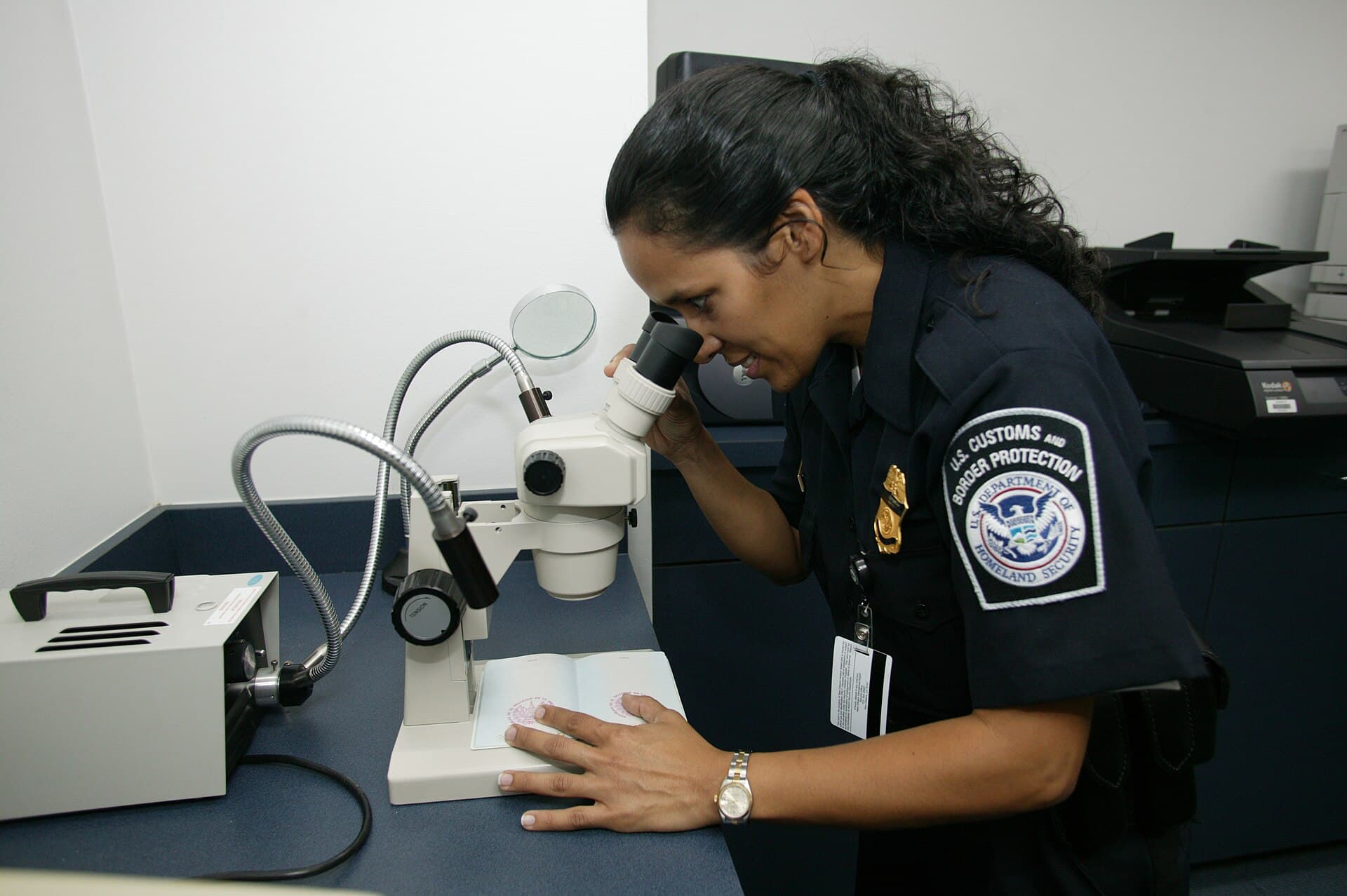 A uniformed U.S. Customs and Border Protection officer leans into a stereomicroscope at an inspection desk in an international airport arrivals hall, examining a traveller's passport under bright indoor light.