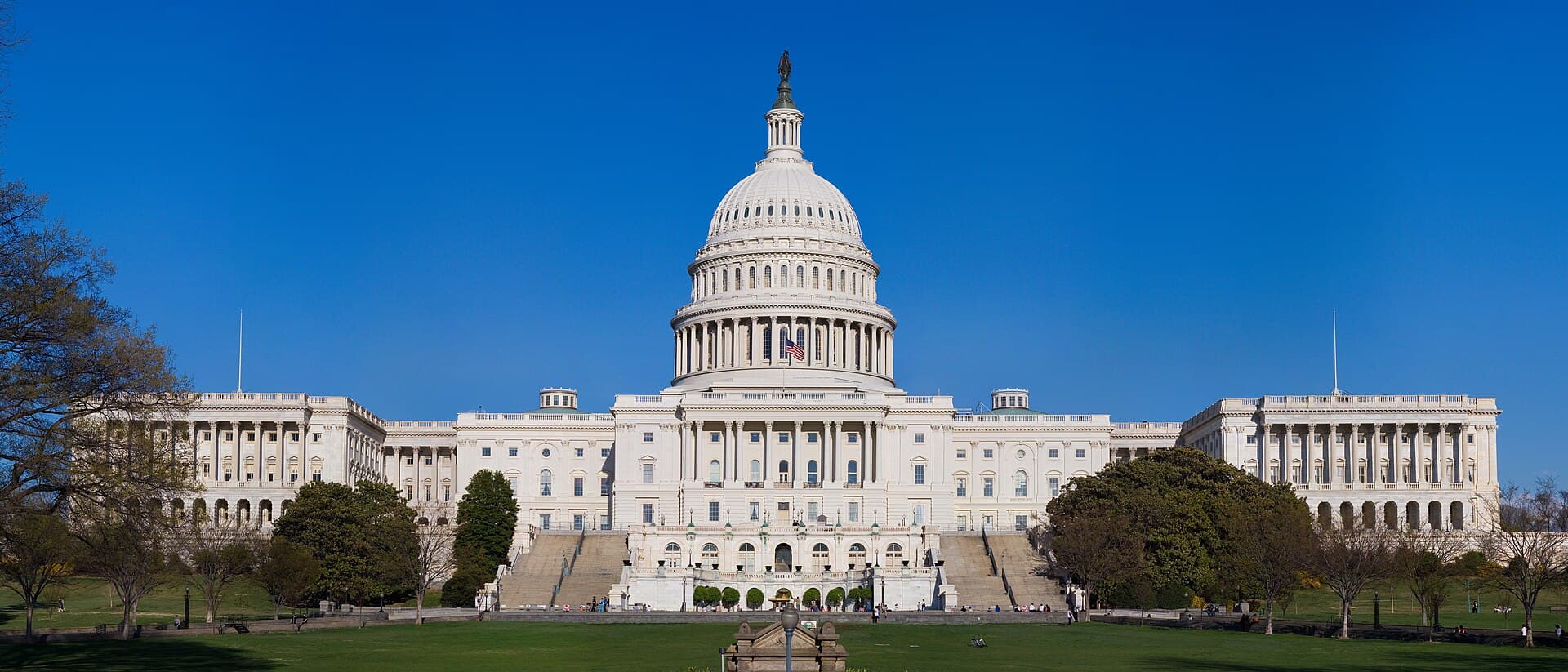 Wide daylight view of the western front of the United States Capitol in Washington, D.C., its central dome and rotunda flanked by the two legislative wings, seen across the terraced steps and lawn under a clear sky.
