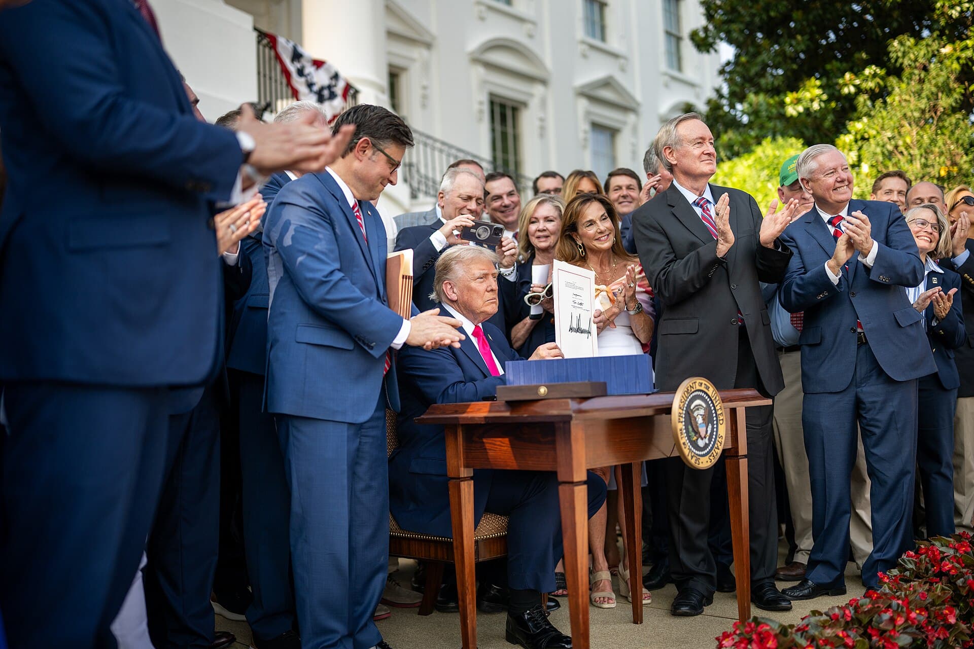 President Donald Trump seated at a desk on the South Lawn of the White House, signing the One Big Beautiful Bill Act in front of onlookers during the 4 July 2025 picnic, in bright afternoon daylight.